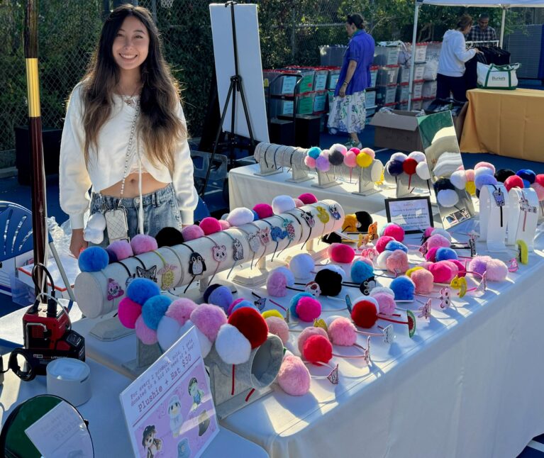 A young woman with long dark hair smiles while standing behind a table at an outdoor market, displaying her handmade pom-pom headband collection. The table is covered with dozens of colorful headbands featuring rhinestone animal charms in a wide variety of pom-pom colors including pink, red, blue, purple, black, and white.