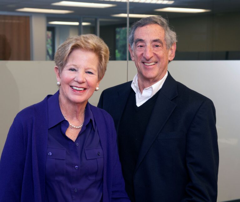 A woman and a man smile together for a professional portrait in an indoor office setting. The woman wears a purple blouse and cardigan with a pearl necklace, and the man wears a black blazer over a white shirt. Both are smiling warmly at the camera.