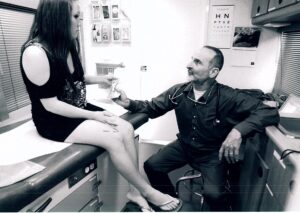 A black-and-white photo of a physician with a stethoscope seated on a stool inside a mobile clinic, speaking with a young woman seated on an exam table.