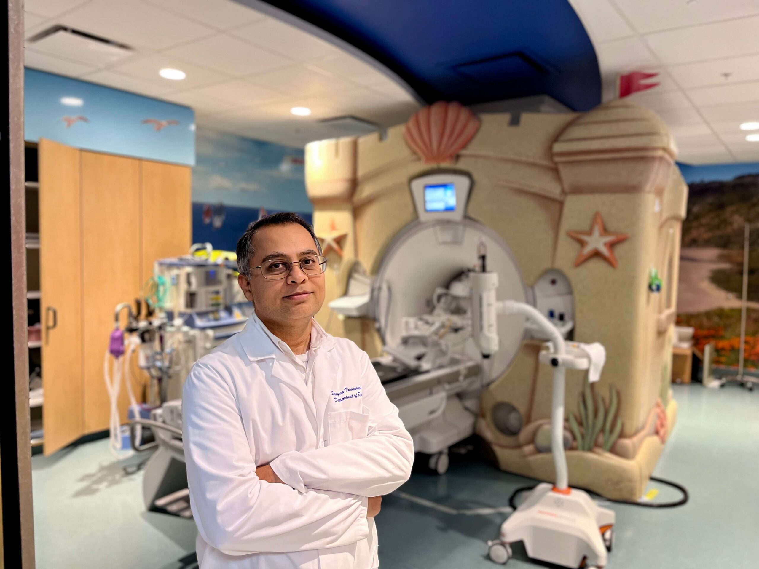 Alt text: A physician in a white lab coat stands with arms crossed in front of a pediatric MRI machine decorated to look like a sand castle, with a colorful ocean-themed mural on the walls behind him.