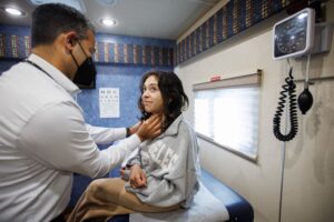 A masked physician examines a teenage girl's neck as she sits on an exam table inside the Teen Van mobile clinic, with a vision chart and blood pressure cuff visible on the wall behind them.