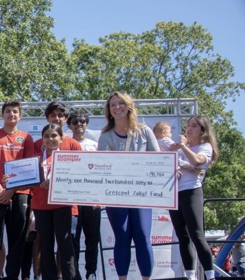 A large group of youth and adults pose on an outdoor stage at the Summer Scamper fundraising event, sponsored by Stanford Medicine. Several participants wear red Summer Scamper t-shirts and medals. One person in a white Summer Scamper hoodie sits on the front edge of the stage. A woman on the right holds a large ceremonial check made out to the Crescent Zakat Fund for $91,266, dated June 21, 2025. Teal balloons and green trees are visible in the background