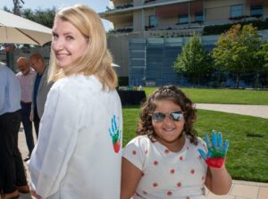 A woman in a white lab coat decorated with a painted handprint looks back over her shoulder and smiles, standing next to a young girl in a strawberry-print outfit and sunglasses who holds up a hand freshly painted in blue, green, and red. They stand outdoors near a table with paint supplies, with a small crowd of people and a lawn in the background.
