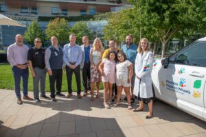 A group of ten adults and two young girls pose outdoors in front of a white car decorated with colorful painted handprints and a 'Hyundai Hope on Wheels — Helping kids fight cancer' logo. The group stands on a paved patio in front of a building with green landscaping, on a sunny day. Several men in business attire stand on the left, a woman in a lab coat covered in painted handprints stands on the right, and two smiling young girls in matching outfits stand in the center front.