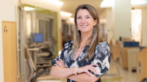 Dr. Alice Bertaina poses with arms crossed and a smile to the camera while standing in the middle of a hospital hallway.
