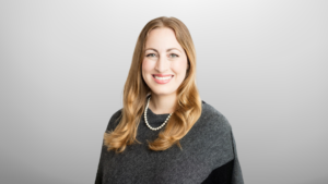 Dr. Michelle Monje wears a dark grey sweater, a necklace and poses and smiles at the camera. Dr. Monje sits in front of a grey backdrop.