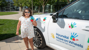 Lucile Packard Children's Hospital Stanford Patient Hero Rubi poses next to the Hyundai Hope on Wheels car while holding up a hand covered in paint. Rubi has just pressed the painted hand on the car as part of the hand painting ceremony.