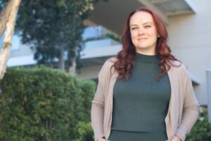 A woman with long, wavy auburn hair smiles gently at the camera. She wears a dark green top with a beige cardigan and stands outdoors in front of greenery and a building with blurred background."