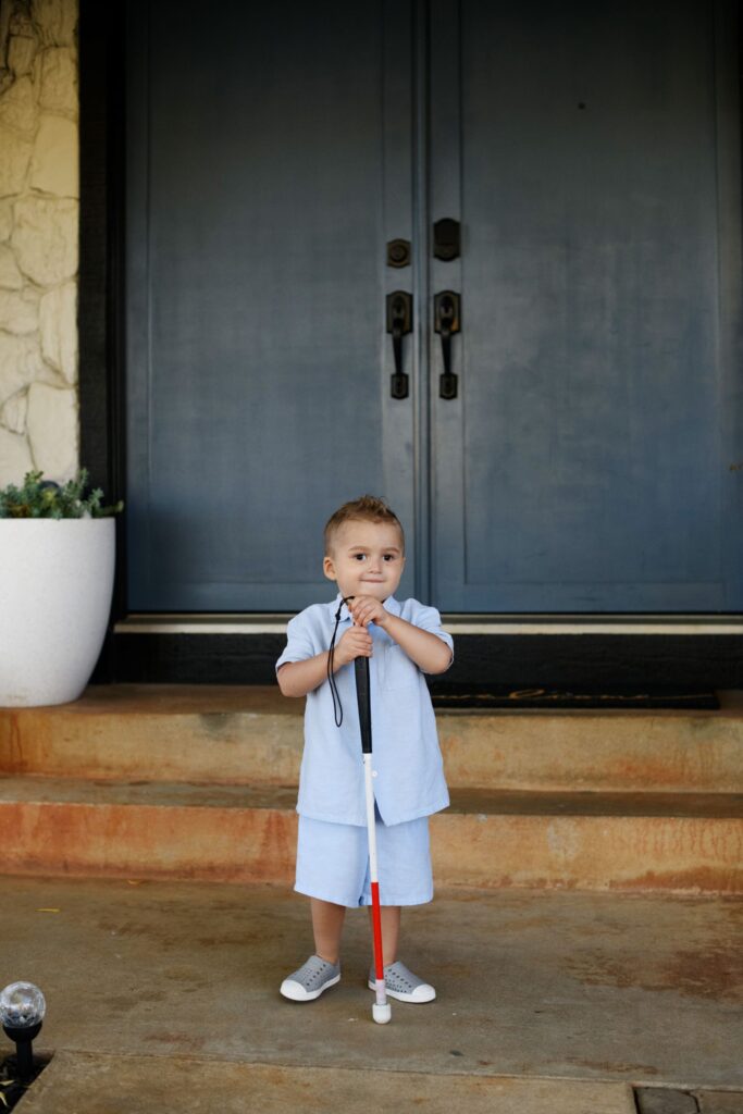 A toddler in a light blue shirt and shorts stands on a front porch in front of a navy double door, holding a white and red mobility cane with both hands and smiling.