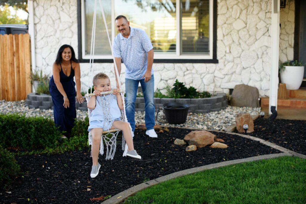 A toddler in light blue clothes swings in a macramé hanging chair in a front yard while a smiling woman and man watch and lean in to push him.