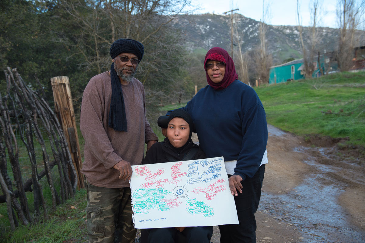 A girl with special health care needs wearing a hijab is seated in her wheelchair outside her home in a rural California town. She and her mother and stepfather are holding her care map.
