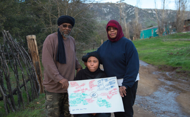 A girl with special health care needs wearing a hijab is seated in her wheelchair outside her home in a rural California town. She and her mother and stepfather are holding her care map.