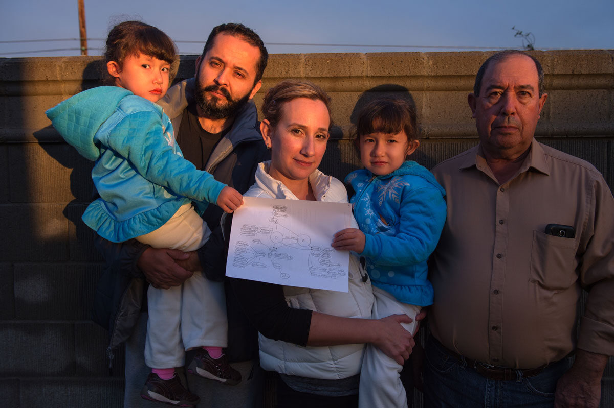 Twin girls with special health care needs are held by their parents while standing next to their grandfather. They are outside, next to a fence, their faces reflecting the sunset. The girls are holding a copy of their care map.