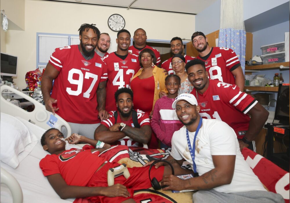Group of football players pose with child in hospital bed.