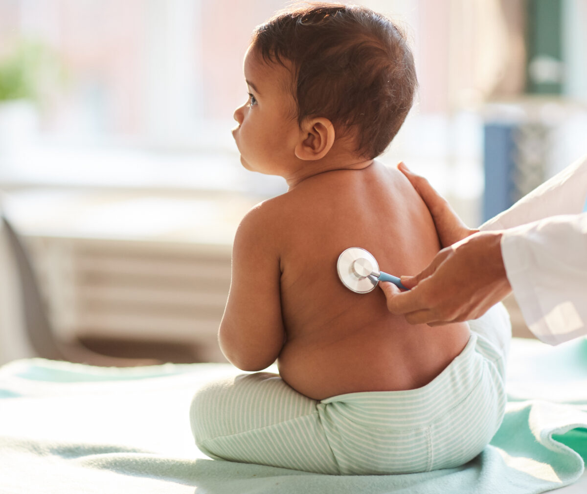 Rear view of shirtless toddler sitting on the table while doctor listening to him with stethoscope