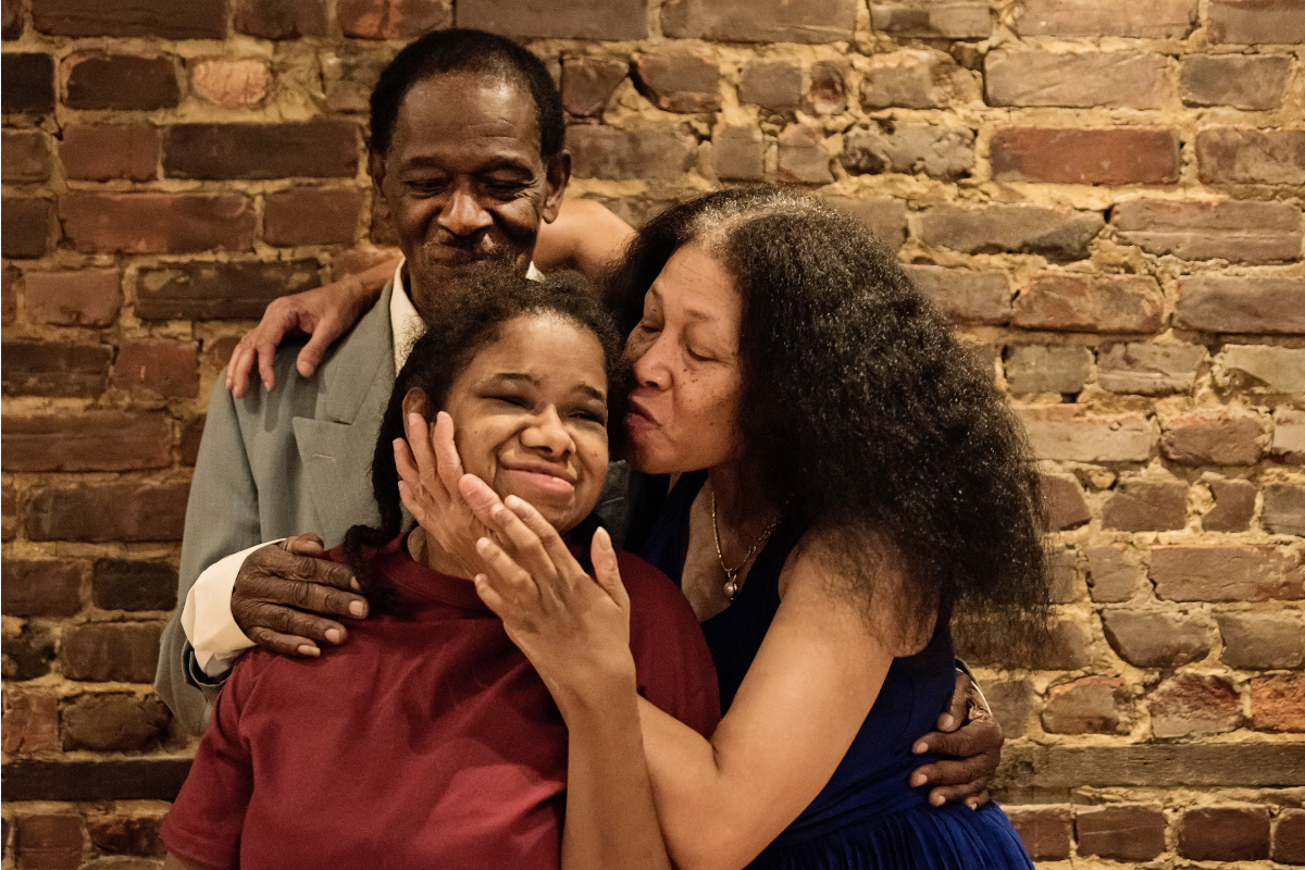An older Black man wearing a gray suit and older Black woman with long hair are embracing a younger Black woman who appears to have developmental disabilities. They are all smiling and standing in front of a brick wall.