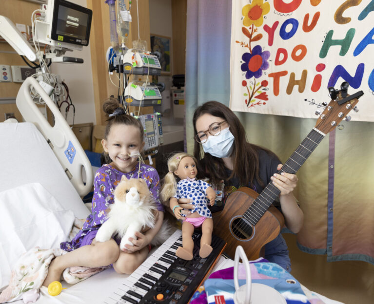 A young patient poses with her musical therapist in her hospital room. There is a keyboard on the bed and the therapist is holding a guitar.
