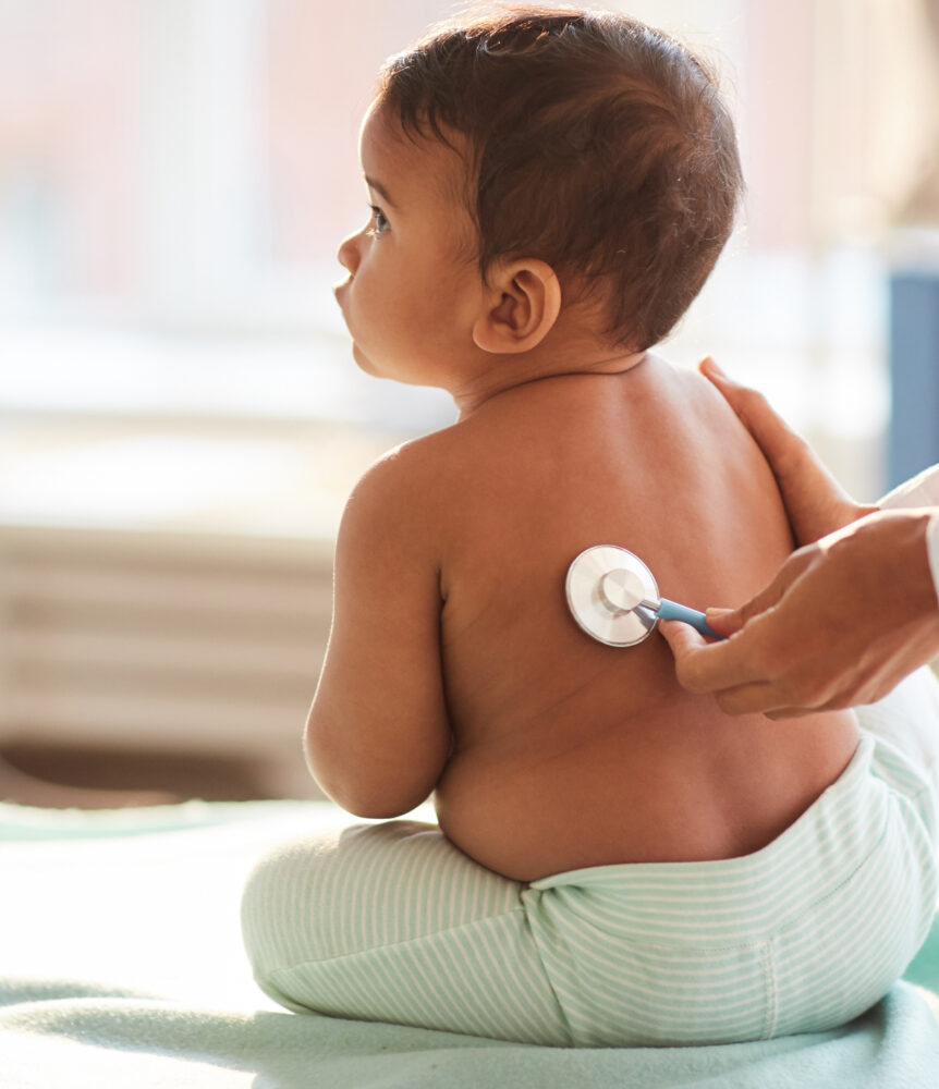 Rear view of shirtless toddler sitting on the table while doctor listening to him with stethoscope