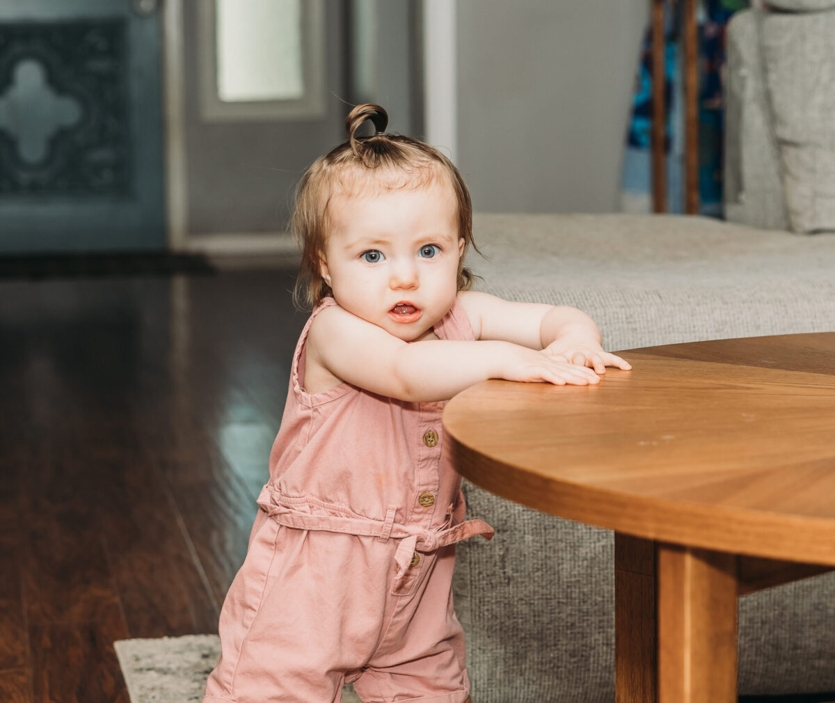 1-year-old girl, Hazel, stands by a table.