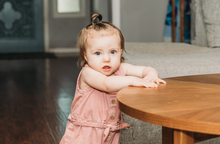 1-year-old girl, Hazel, stands by a table.