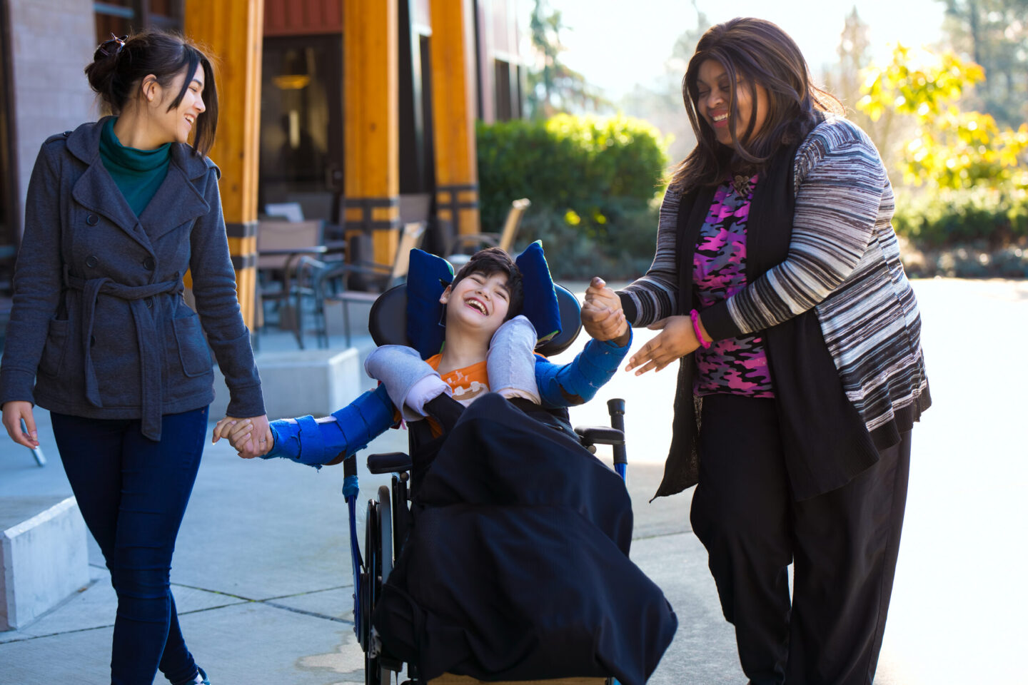 Photo of a child in a wheelchair smiling and holding hands with caregivers while on a walk outdoors.