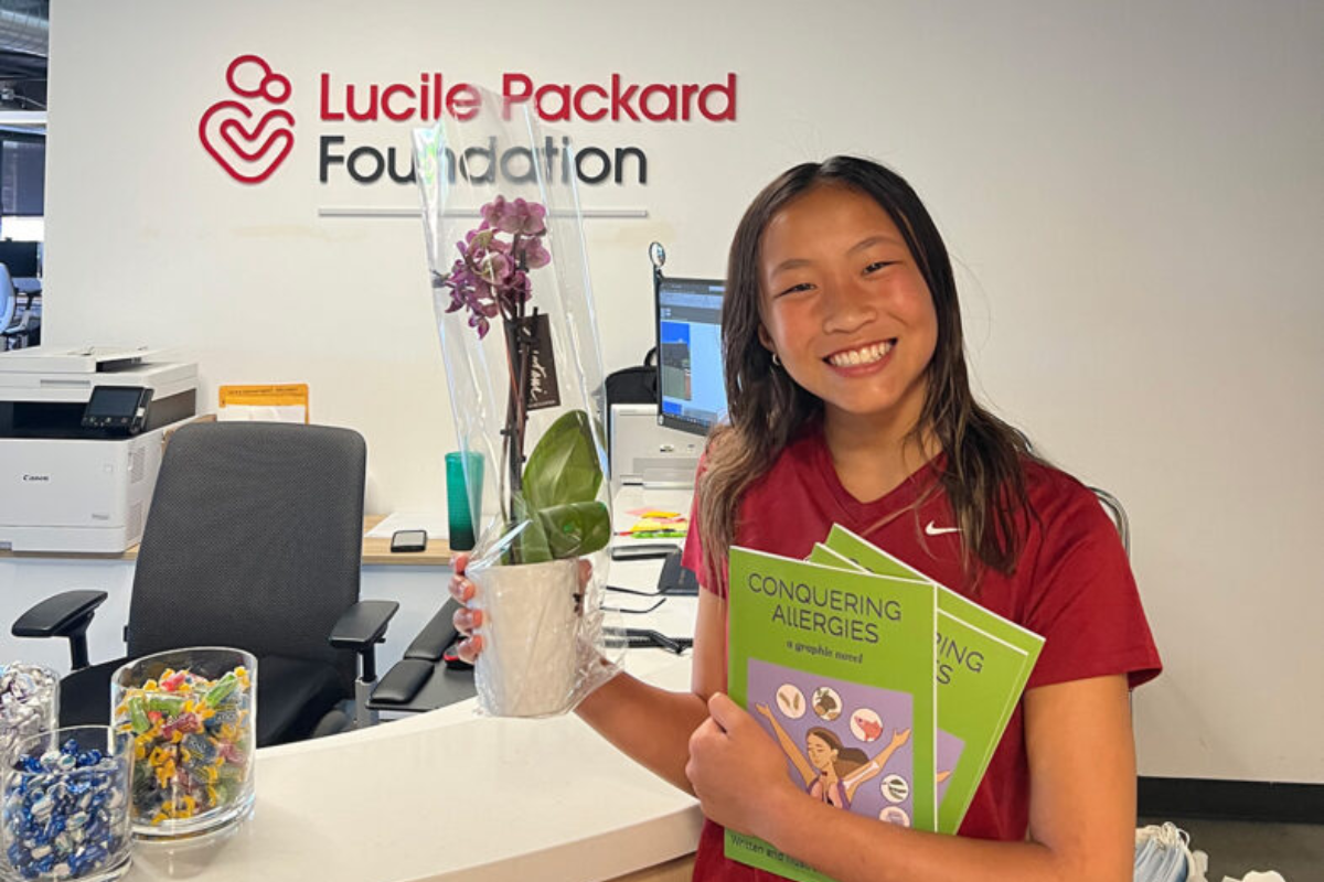 A smiling former Packard Children's allergy patient standing in front of the Lucile Packard Foundation for Children's Health logo at the Foundation's office.
