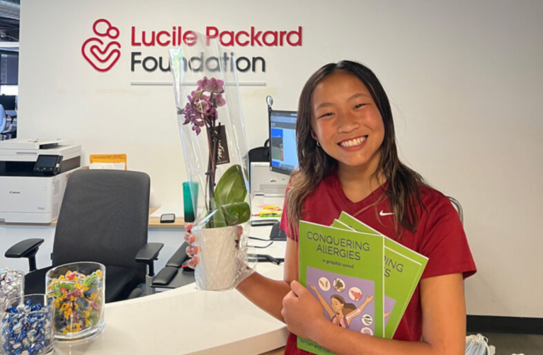 A smiling former Packard Children's allergy patient standing in front of the Lucile Packard Foundation for Children's Health logo at the Foundation's office.