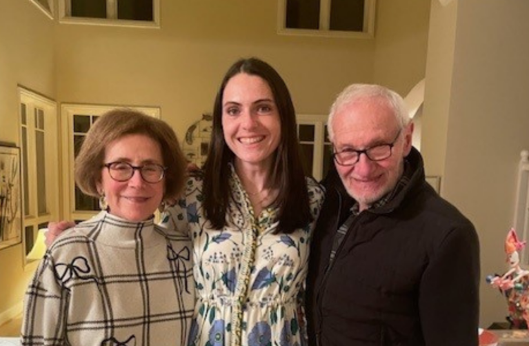 Three adults stand side by side indoors, smiling at the camera; a former packard patient in a patterned dress stands between their grandparents.