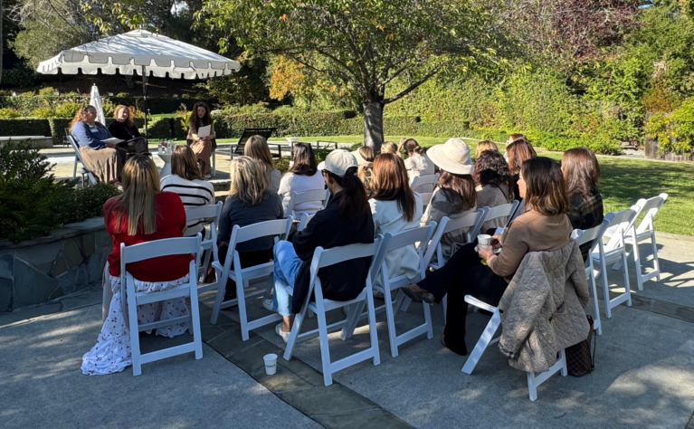 Ambassador members sitting in chairs outside in a sunny garden listening to speakers that are sitting on a stage.