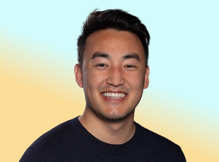 Smiling person in a dark sweater facing the camera in a studio-style headshot against a soft blue and yellow gradient background.
