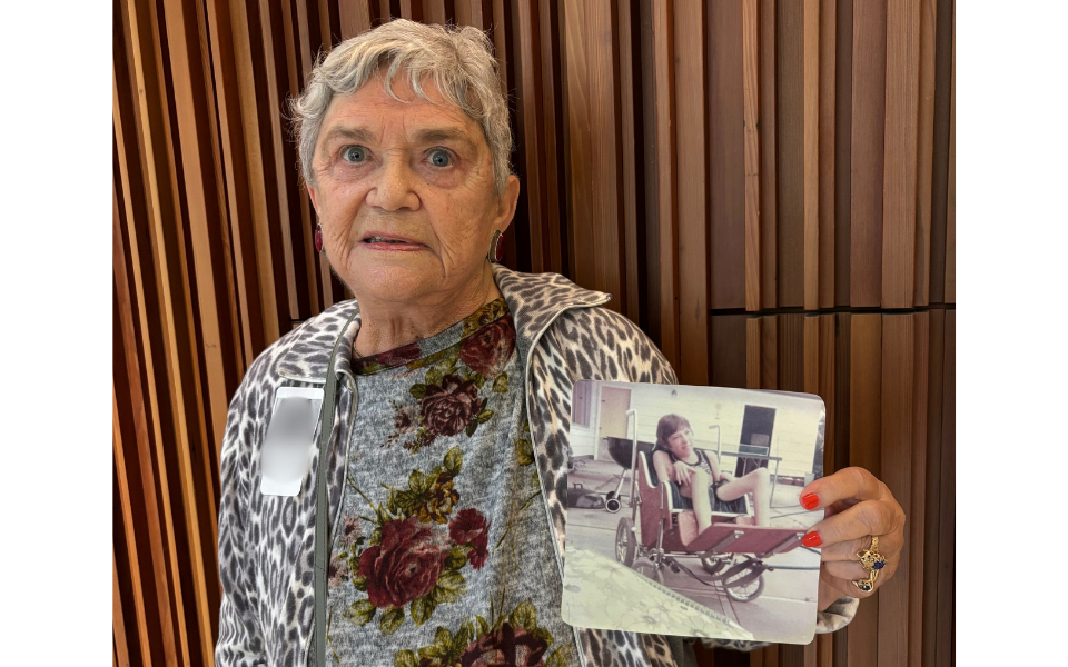 An older adult smiles while holding up a printed photograph of their late child in their right hand, presenting it toward the camera. The focus is on the photograph and the person’s expression, capturing a moment of remembrance and connection.