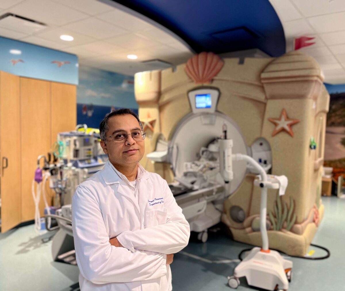 Alt text: A physician in a white lab coat stands with arms crossed in front of a pediatric MRI machine decorated to look like a sand castle, with a colorful ocean-themed mural on the walls behind him.