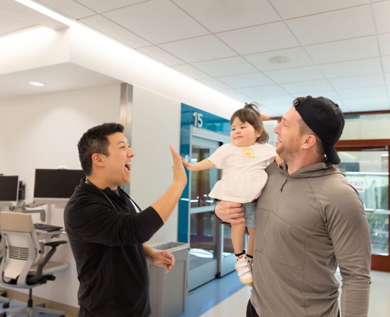 A clinician shares a high five with Audrey, a smiling toddler being held by her father, in a bright hospital hallway at Lucile Packard Children's Hospital Stanford.