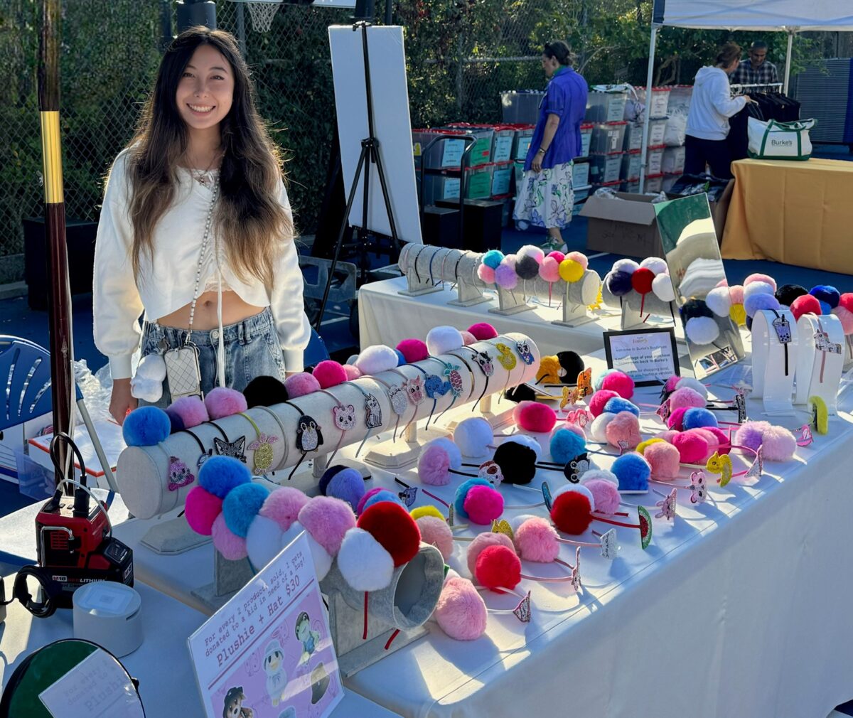 A young woman with long dark hair smiles while standing behind a table at an outdoor market, displaying her handmade pom-pom headband collection. The table is covered with dozens of colorful headbands featuring rhinestone animal charms in a wide variety of pom-pom colors including pink, red, blue, purple, black, and white.