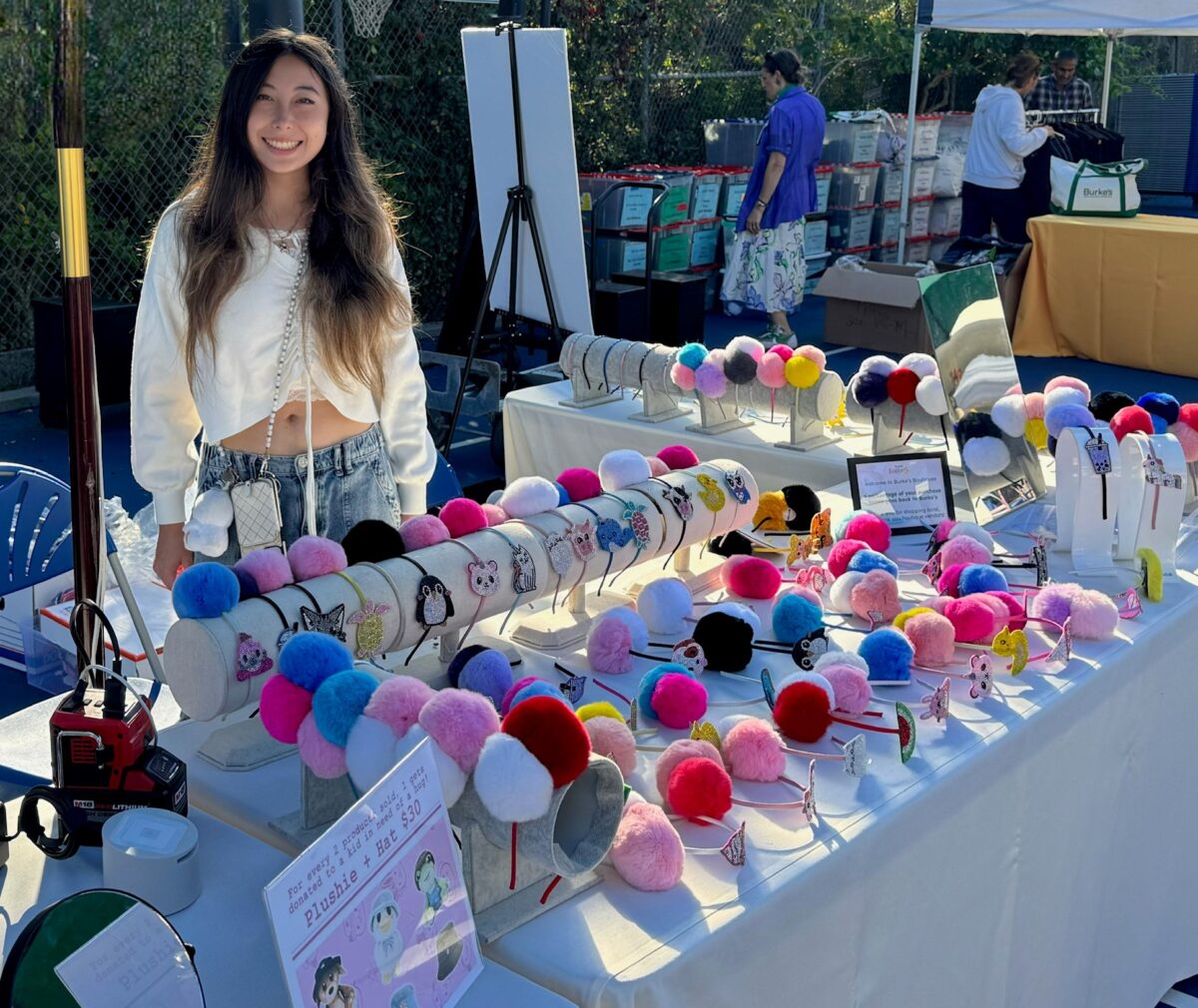 A young woman with long dark hair smiles while standing behind a table at an outdoor market, displaying her handmade pom-pom headband collection. The table is covered with dozens of colorful headbands featuring rhinestone animal charms in a wide variety of pom-pom colors including pink, red, blue, purple, black, and white.
