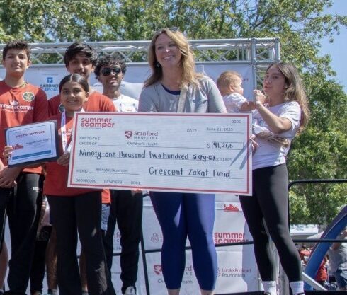 A large group of youth and adults pose on an outdoor stage at the Summer Scamper fundraising event, sponsored by Stanford Medicine. Several participants wear red Summer Scamper t-shirts and medals. One person in a white Summer Scamper hoodie sits on the front edge of the stage. A woman on the right holds a large ceremonial check made out to the Crescent Zakat Fund for $91,266, dated June 21, 2025. Teal balloons and green trees are visible in the background
