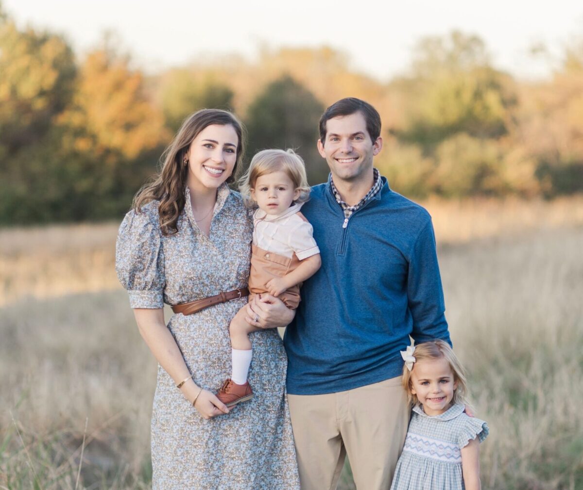 A family of four poses together in an open field during golden hour, with autumn trees in the background.