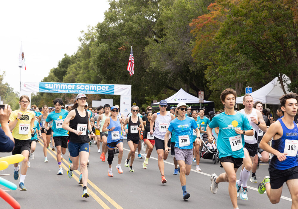 An action shot of a dense pack of runners coming toward the camera on a tree-lined road under the Summer Scamper event banner, with the lead runner wearing a 'MUSTANGS' single
