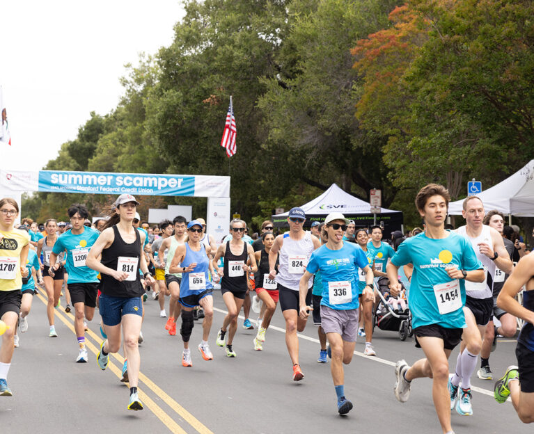An action shot of a dense pack of runners coming toward the camera on a tree-lined road under the Summer Scamper event banner, with the lead runner wearing a 'MUSTANGS' single