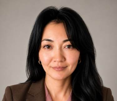 Professional headshot of a woman in business attire against a neutral gray background, wearing a brown blazer over a dusty rose blouse, with shoulder-length black hair, gold hoop earrings, and a composed, approachable expression.