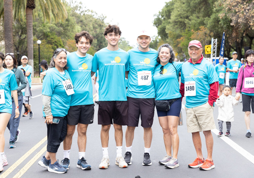 A multigenerational family of six in matching light-blue 'summer scamper' t-shirts posing on a closed road during the race