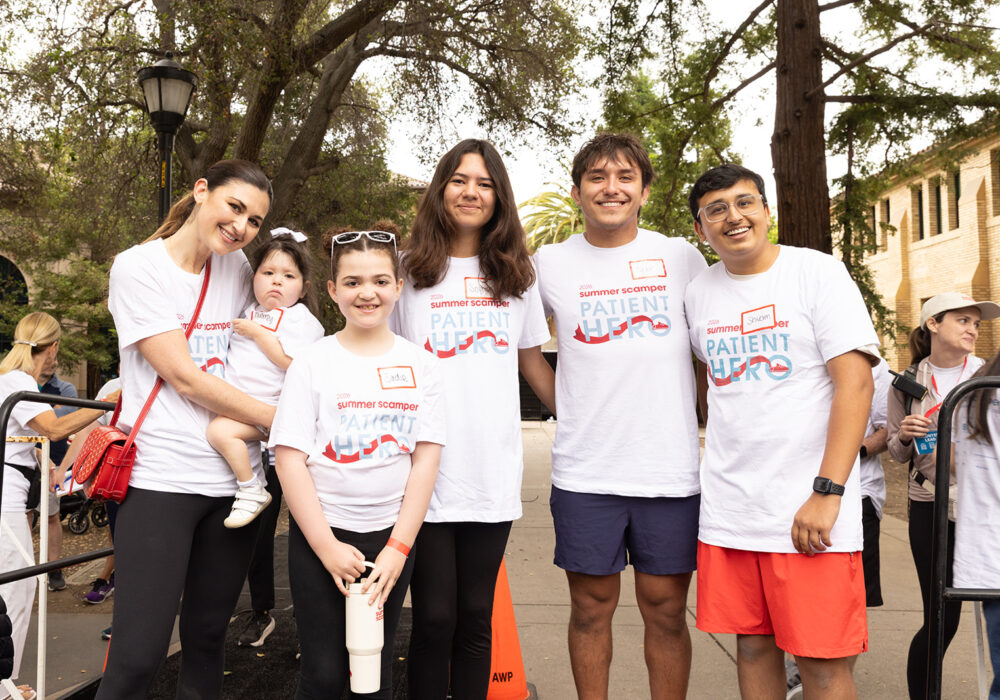 Six individuals wearing white '2026 Summer Scamper Patient Hero' t-shirts with handwritten name tags, including a woman holding a toddler, posing on a paved path with Spanish-style architecture in the background
