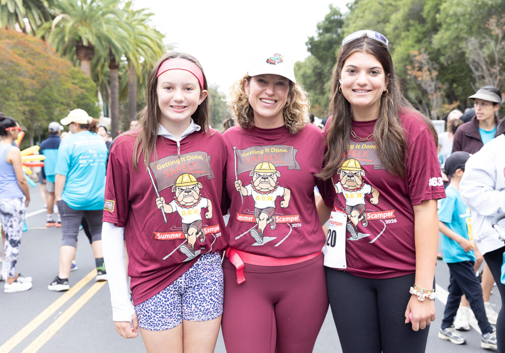 Three women in matching maroon 'Getting It Done, SAFELY. Summer Scamper 2026' t-shirts featuring a cartoon bulldog mascot posing on the race road;