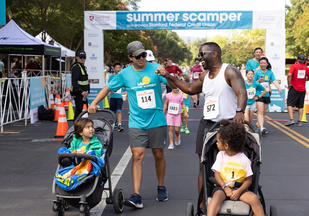 Two fathers pushing strollers and sharing a fist bump as they walk/run the course near the start/finish line banner, with other participants visible in the background.