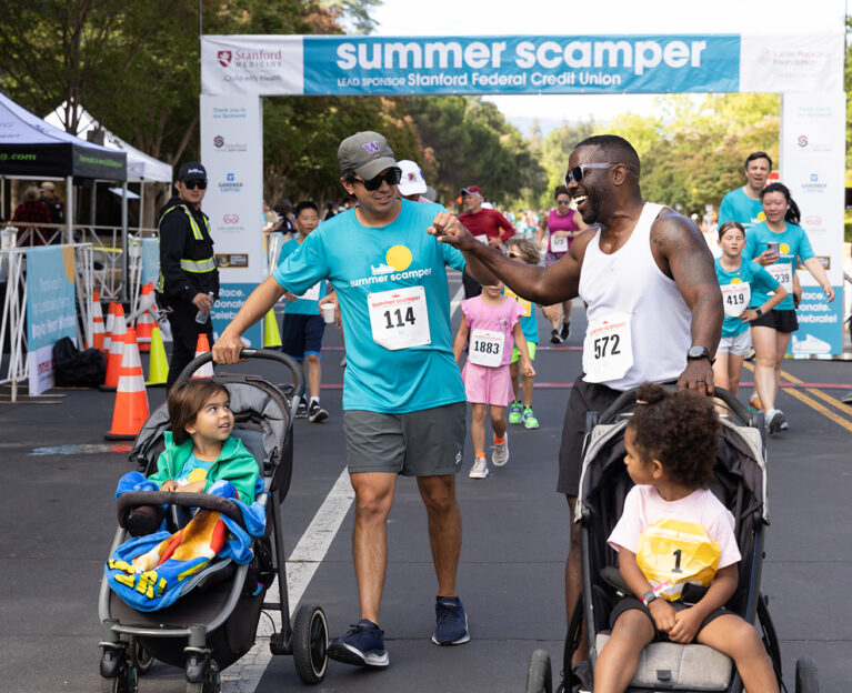 Two fathers pushing strollers and sharing a fist bump as they walk/run the course near the start/finish line banner, with other participants visible in the background.