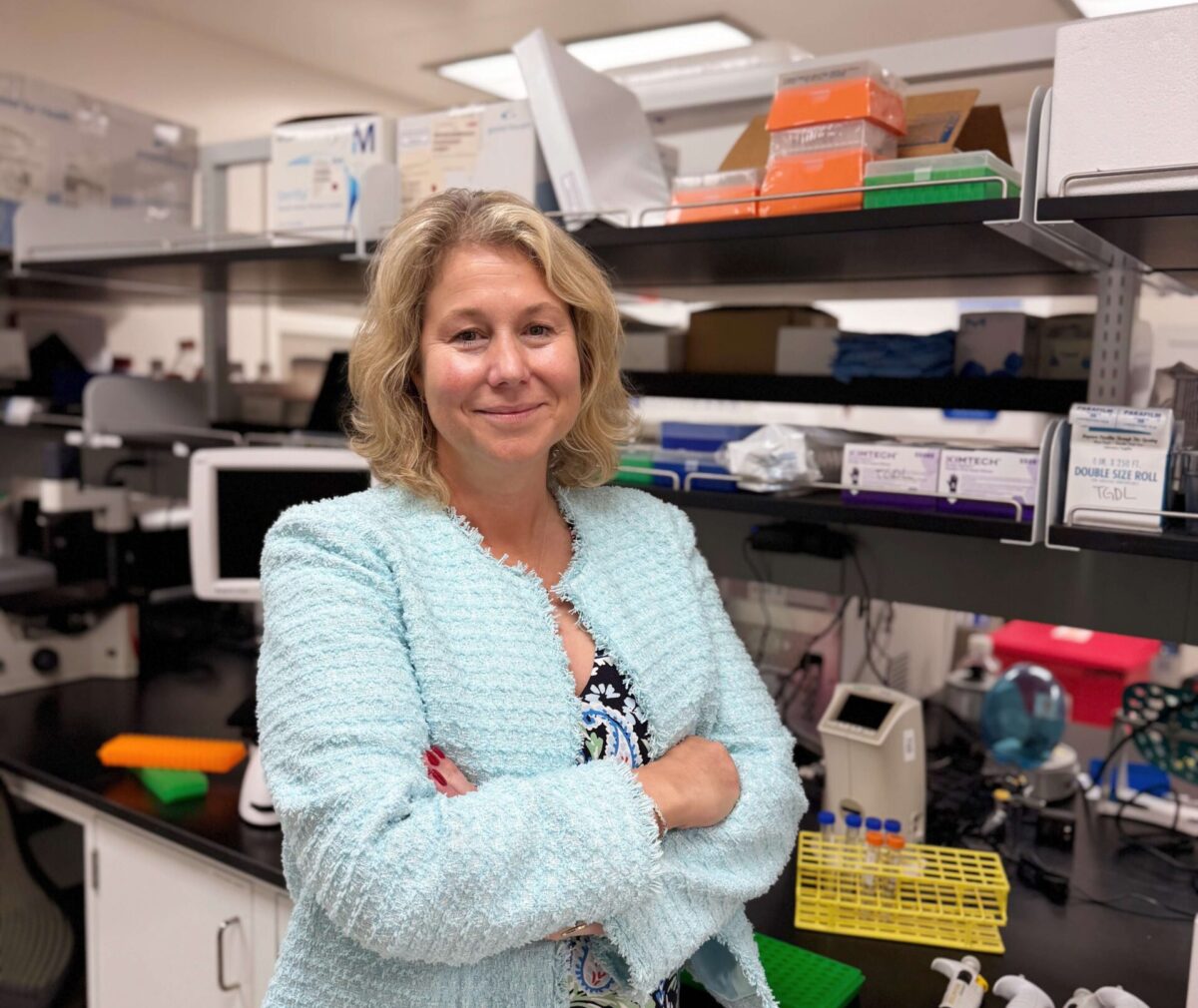 A woman with wavy shoulder-length blonde hair stands with arms crossed in a laboratory, smiling at the camera. She wears a light teal textured cardigan over a dark floral paisley top and skirt. Behind her, lab shelves hold pipette tip boxes, Kimtech wipes, and other supplies, while the black countertop displays a yellow test tube rack, several white multichannel pipettes, and assorted lab equipment.