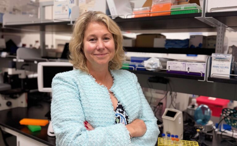 A woman with wavy shoulder-length blonde hair stands with arms crossed in a laboratory, smiling at the camera. She wears a light teal textured cardigan over a dark floral paisley top and skirt. Behind her, lab shelves hold pipette tip boxes, Kimtech wipes, and other supplies, while the black countertop displays a yellow test tube rack, several white multichannel pipettes, and assorted lab equipment.