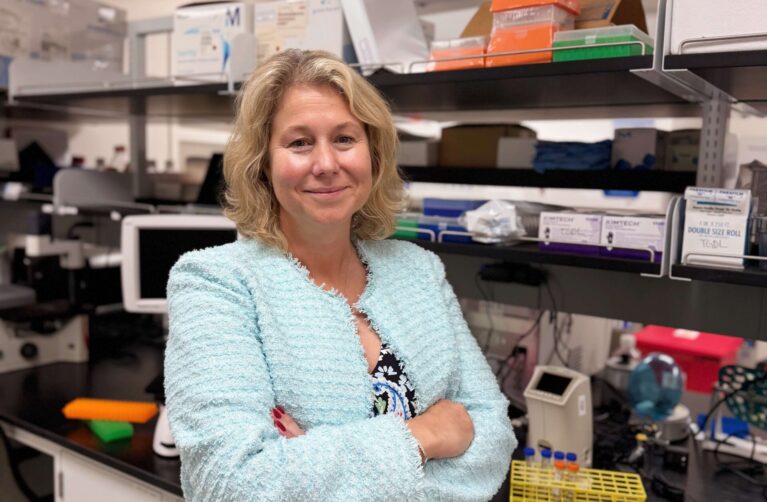 A woman with wavy shoulder-length blonde hair stands with arms crossed in a laboratory, smiling at the camera. She wears a light teal textured cardigan over a dark floral paisley top and skirt. Behind her, lab shelves hold pipette tip boxes, Kimtech wipes, and other supplies, while the black countertop displays a yellow test tube rack, several white multichannel pipettes, and assorted lab equipment.