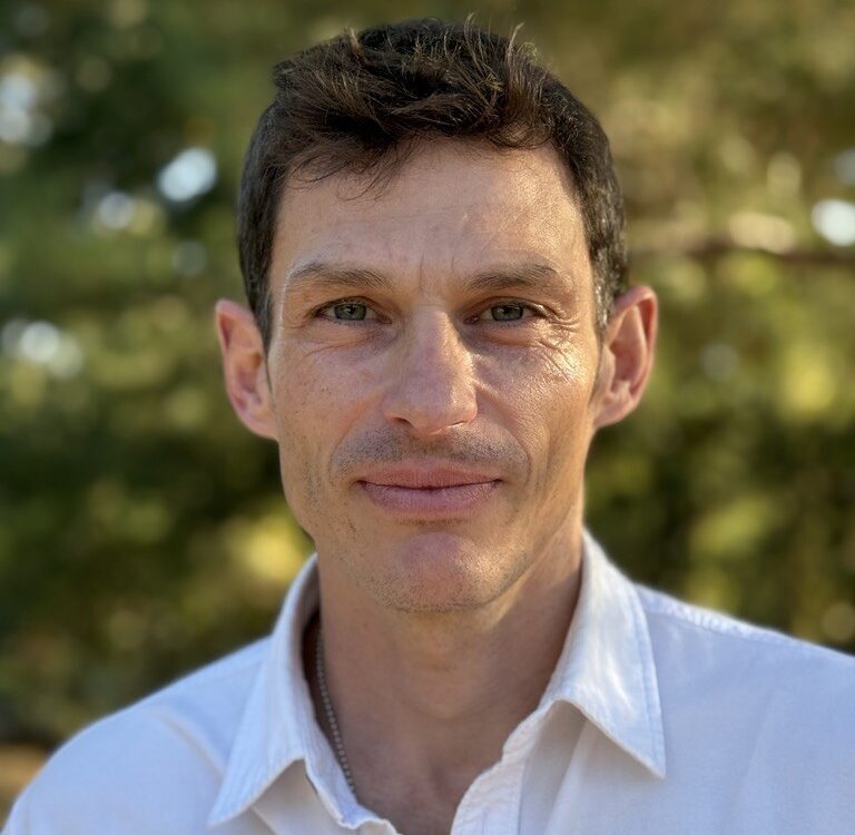 Professional headshot of a man with short, wavy dark brown hair, smiling warmly at the camera. He has fair skin, green eyes, and light stubble, and is wearing a white button-down collared shirt with a thin necklace visible at the neckline. The background is a softly blurred outdoor scene of green foliage, suggesting natural daylight.