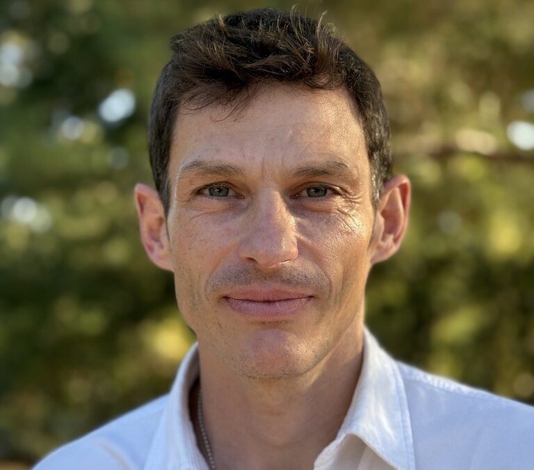 Professional headshot of a man with short, wavy dark brown hair, smiling warmly at the camera. He has fair skin, green eyes, and light stubble, and is wearing a white button-down collared shirt with a thin necklace visible at the neckline. The background is a softly blurred outdoor scene of green foliage, suggesting natural daylight.